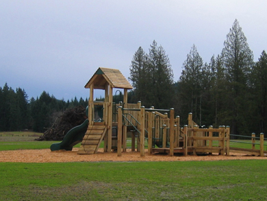 A new wooden play structure on sand, in a grassy area of the new community park
