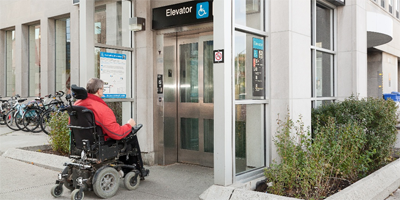 A person using a powered wheelchair approaches an accessible elevator entrance at a transit station, marked with wheelchair accessibility signage.