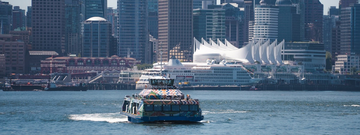 A ferry with detailed Indigenous artwork moves across calm water. In the background, a city skyline with modern high-rise buildings and a unique waterfront structure sits under a bright blue sky.