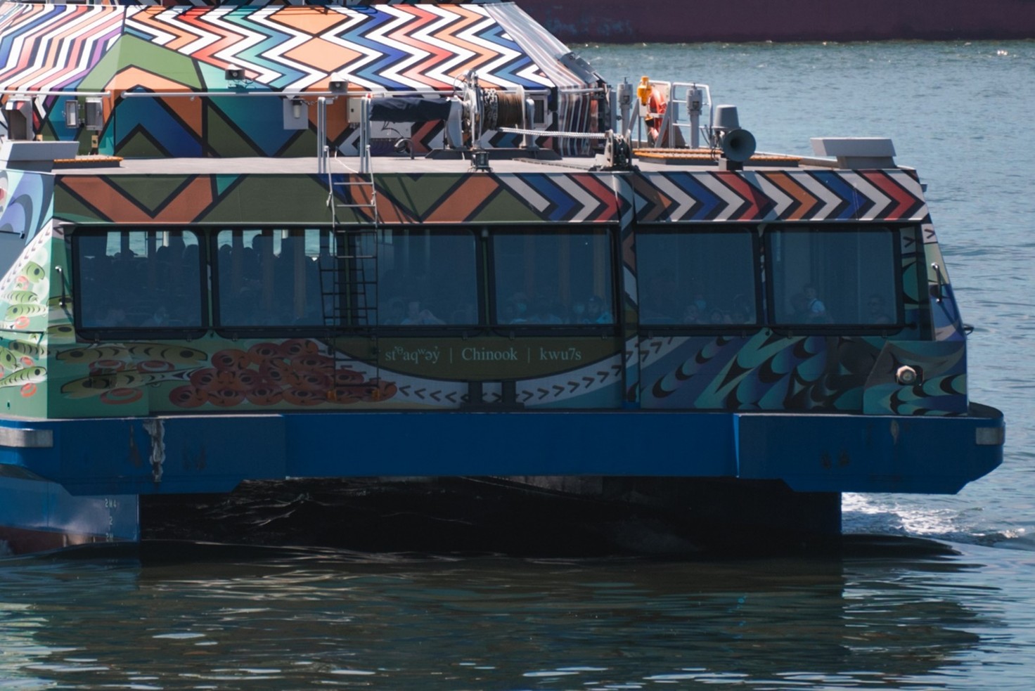 A close-up view of a ferry on top of blue water, decorated with vibrant Indigenous artwork featuring intricate geometric patterns and depictions of natural elements.