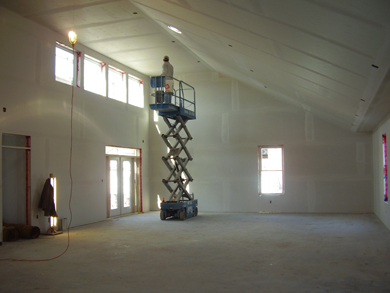 A construction worker uses an electric lift truck to install sheet rock and tape the interior walls and ceiling