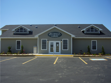 The front of the new Weymouth Public Library building, with grey siding exterior walls and a paved parking lot