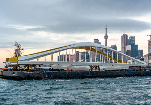 A barge carrying a modern bridge across the water, with the city of Toronto skyline in the background.