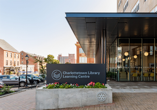 Exterior of a library with floor to ceiling windows, a sign next to the entrance with the text “Charlottetown Library Learning Centre”.