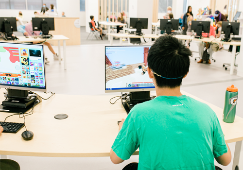 A teen at a computer station, playing a video game. Other computers and people are in the background of a spacious library.