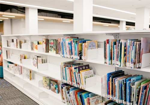 A neatly organized children’s section in a library with low white bookshelves filled with colorful books.