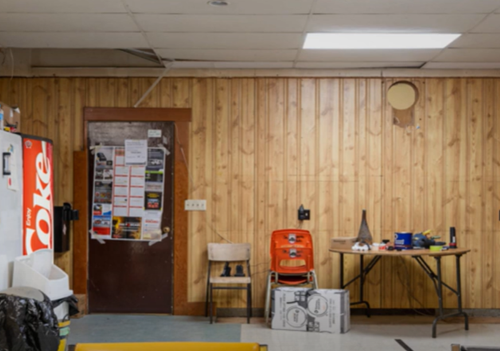 BEFORE: Interior of fire hall. Wood paneling on all walls. Long table with chair. Various item scattered throughout room. 