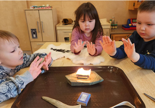 Three children around a table with their hands reached out around a small flame on a tray.