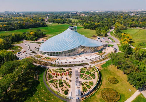 An aerial view of a large building with a glass roof, surrounded by gardens and greenspace.