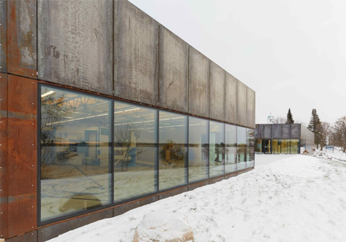 A snowy wooden walkway leading to a modern metal building with large windows.